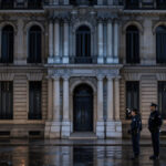 Façade nocturne de la préfecture de police de Paris, éclairée par une lumière froide, avec deux agents en uniforme flous à l’arrière-plan et des reflets sur le sol humide.